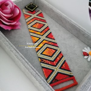 Può includere: Un braccialetto di perline rosse, arancioni, gialle, nere e dorate con un motivo geometrico. Il braccialetto è in una scatola grigia.