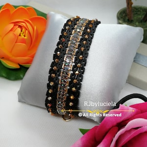 以下が含まれることがあります： 金色のアクセントと透明なクリスタルが付いた黒いビーズのブレスレット。ブレスレットは、ピンクのバラが前景にある白い枕の上に置かれています。