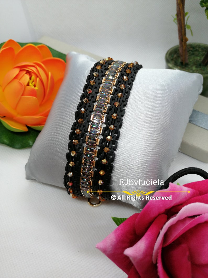 以下が含まれることがあります： 金色のアクセントと透明なクリスタルが付いた黒いビーズのブレスレット。ブレスレットは、ピンクのバラが前景にある白い枕の上に置かれています。