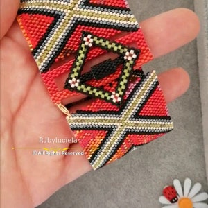 以下が含まれることがあります： 赤、黒、白、黄色の幾何学模様が特徴の手作りのビーズブレスレット。ブレスレットは正方形で、中央に正方形のデザインがあります。ブレスレットは手で持たれています。