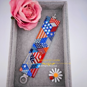 May include: A beaded bracelet with a colorful geometric pattern featuring red, white, and blue colors. The bracelet has a silver clasp and is displayed in a gray velvet box. The text "Renovatio By Luciela" and "All Rights Reserved" is visible on the box.