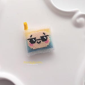 Könnte beinhalten: Ein quadratischer Perlenanhänger mit einem verpixelten Charaktergesicht mit schwarzer Sonnenbrille, rosa Wangen und blondem Haar, gekrönt von einem kleinen gelben Akzent. Das Design besteht aus einem Raster kleiner Perlen mit einer transparenten Schicht.