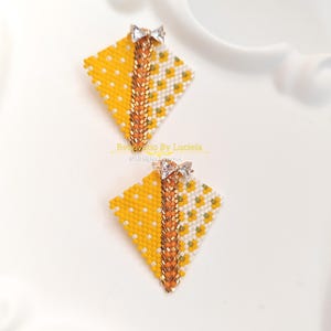 May include: A pair of handmade beaded earrings. Each earring is a yellow and white diamond shape with a decorative chain and a small bow at the top. The earrings have a floral pattern and are on a white surface.