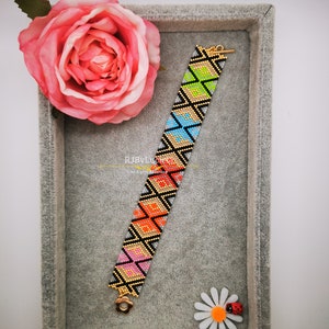 Può includere: Un braccialetto colorato con perline e un motivo geometrico in tonalità di rosa, arancio, giallo, verde, blu e nero. Il braccialetto ha chiusure dorate ed è esposto in una scatola di velluto grigio.