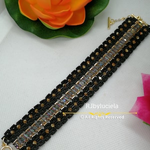 Può includere: Un braccialetto nero con dettagli dorati e pietre rettangolari iridescenti. Il braccialetto ha una chiusura a scorrimento e  è composto da più file di perline.