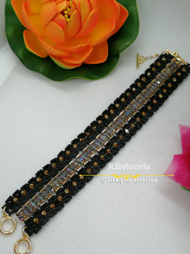 以下が含まれることがあります： ゴールドのアクセントと虹色の長方形の石が付いた黒いブレスレット。ブレスレットはトグル留め金で、複数のビーズの列でできています。