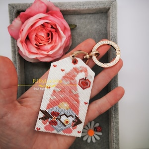 May include: A pink and white beaded gnome keychain with a red heart and a red apple. The keychain is attached to a gold-colored metal ring with clear rhinestones.