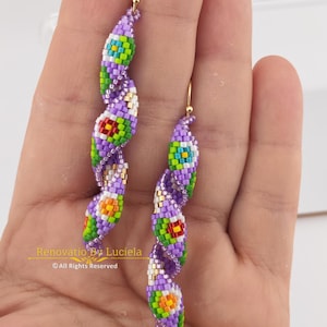 Può includere: Un paio di orecchini di perline con un design a spirale. Gli orecchini presentano una combinazione di perline viola, verdi, gialle, rosse e bianche. Gli orecchini hanno ganci color oro. Il testo "Renovatio By Luciela" e "All Rights Reserved" è visibile.