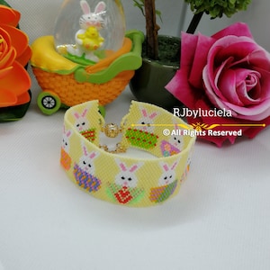 Op de afbeelding: Een kralenarmband met een gele achtergrond en een herhalend paashaaspatroon. De armband heeft een gouden sluiting en staat tegen een achtergrond van bloemen en een decoratieve sneeuwbol met Pasen als thema.