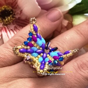 May include: A beaded butterfly ring with purple, blue, and yellow beads. The ring is on a finger and the background is a blurry image of a flower.