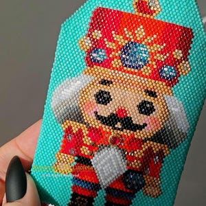 May include: A beaded ornament featuring a nutcracker in a red and gold uniform with a blue crown. The ornament is on a turquoise background. The text "Removable by Luciela" is at the bottom of the ornament.