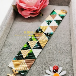 Può includere: Un braccialetto di perline con un motivo geometrico di triangoli verdi, bianchi, dorati e marroni. Il braccialetto ha una chiusura dorata ed è mostrato su una superficie grigia con una rosa rosa e una margherita bianca.