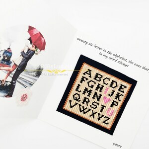 May include: A white card with a black border and a cross-stitch design of the alphabet with a pink heart in the middle. The text on the card reads "twenty six letter in the alphabet, the ones that in my mind always" and "yours".