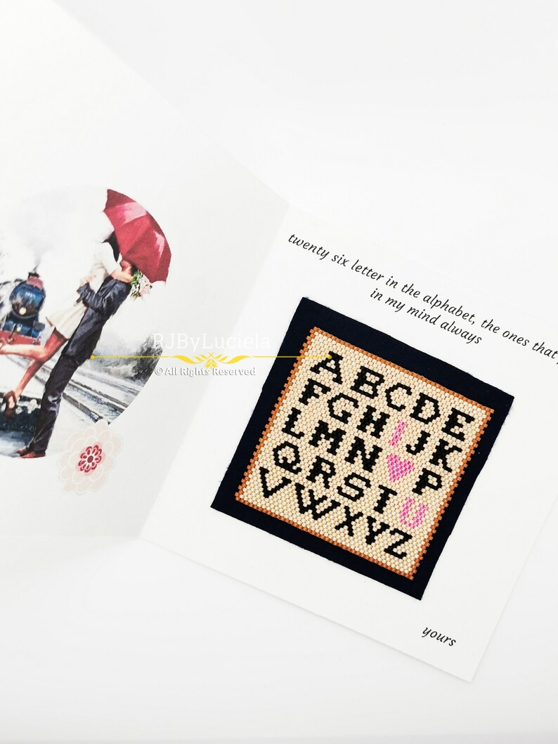 May include: A white card with a black border and a cross-stitch design of the alphabet with a pink heart in the middle. The text on the card reads "twenty six letter in the alphabet, the ones that in my mind always" and "yours".
