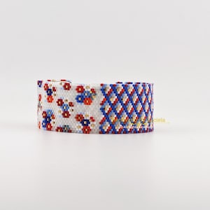 May include: A wide beaded bracelet with two contrasting designs. One side has a white base with red, blue, and yellow floral patterns. The other side features a geometric diamond pattern in red, white, and blue. The bracelet is likely handmade.