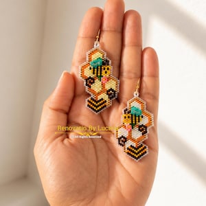 Puede incluir: Pendientes hechos a mano con cuentas en un diseño de panal, con un motivo de abeja. Los pendientes son principalmente amarillos, negros y blancos, con toques de turquesa y rosa. Los pendientes se sostienen en una mano. El texto "Renovatio By Luciela" es visible.