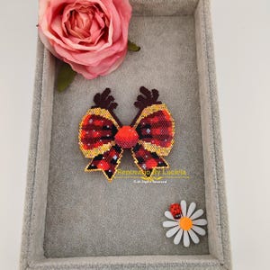 Puede incluir: Un lazo de cuentas rojo y dorado con un centro rojo, en una caja forrada de terciopelo gris. El lazo está decorado con pequeñas cuentas blancas. El texto "Renovatio By Luciela © All Rights Reserved" está impreso debajo del lazo.