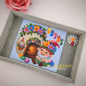 Puede incluir: Un patrón de punto de cruz colorido de un gnomo sentado frente a una pequeña casa decorada con flores y huevos de Pascua. El gnomo lleva un sombrero verde con un pompón rojo y una corona de flores. La casa tiene una puerta marrón y una ventana. El patrón está rodeado de un marco gris.