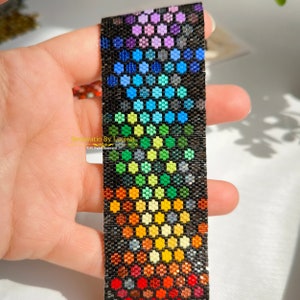 May include: A rainbow-coloured beaded bracelet with a black background. The beads are arranged in a hexagonal pattern, creating a geometric design. The bracelet is made with small, colourful beads.