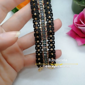 以下が含まれることがあります： 虹色の長方形の石が並んだ黒と金のブレスレット。ブレスレットは小さな黒と金のビーズで作られています。