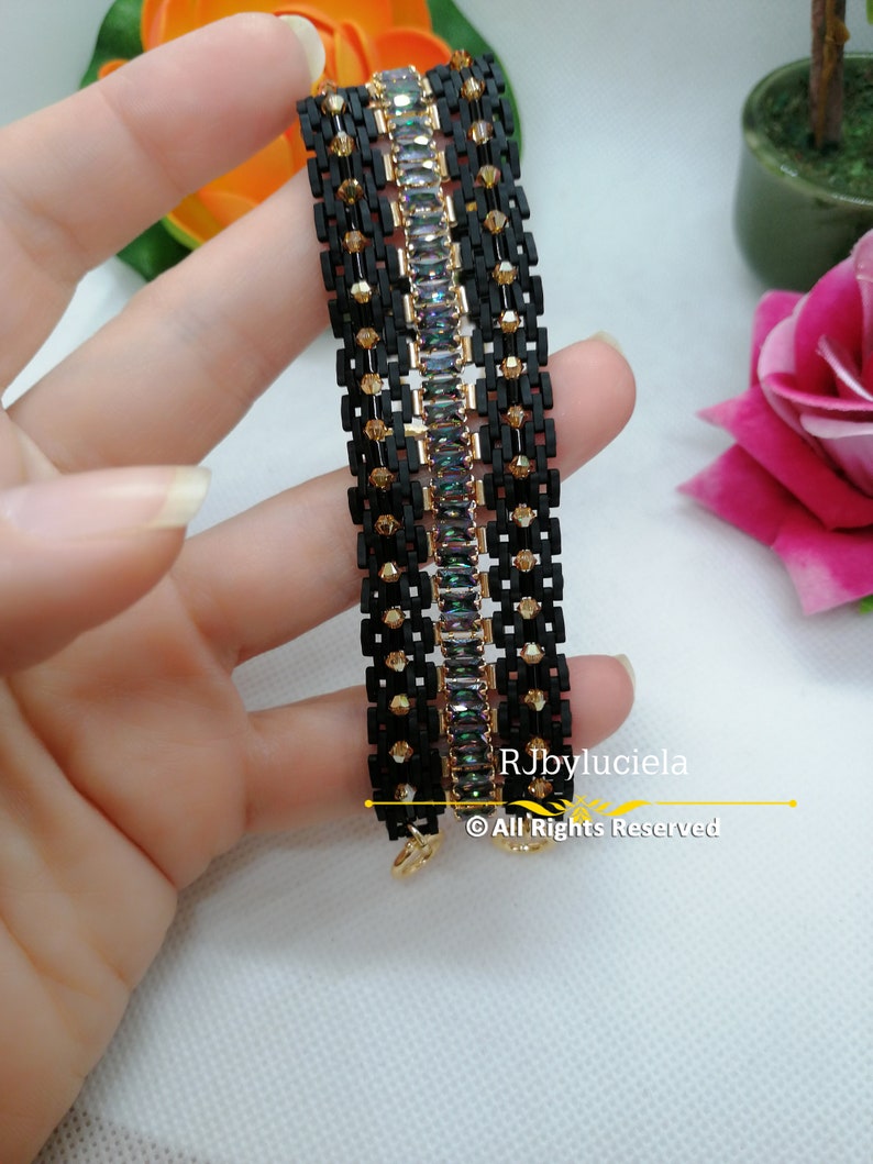 以下が含まれることがあります： 虹色の長方形の石が並んだ黒と金のブレスレット。ブレスレットは小さな黒と金のビーズで作られています。