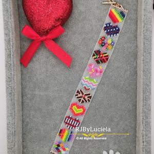 Può includere: Un braccialetto di perline con sfondo bianco e disegni di cuori colorati. Il braccialetto ha una chiusura dorata ed è esposto in una scatola di velluto grigio con un cuore rosso e un fiocco.