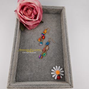 K&ouml;nnte beinhalten: Bunte Perlenohrringe mit goldfarbenen Haken liegen auf einem grauen Samttablett, neben einer rosa Rose und einer G&auml;nsebl&uuml;mchen mit einem Marienk&auml;fer. Die Ohrringe haben ein geometrisches Design in Regenbogenfarben. Der Text "Renovatio By Luciela" ist sichtbar.