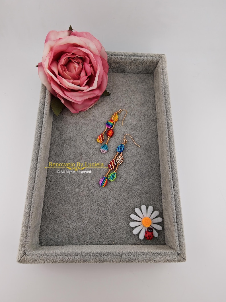 K&ouml;nnte beinhalten: Bunte Perlenohrringe mit goldfarbenen Haken liegen auf einem grauen Samttablett, neben einer rosa Rose und einer G&auml;nsebl&uuml;mchen mit einem Marienk&auml;fer. Die Ohrringe haben ein geometrisches Design in Regenbogenfarben. Der Text "Renovatio By Luciela" ist sichtbar.