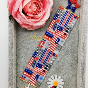 May include: A beaded bracelet with a red, white, and blue American flag pattern. The bracelet is in a gray box with a pink rose and a white daisy with a red ladybug.