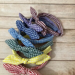 May include: A stack of six gingham fabric headbands in various colors, including red, yellow, blue, green, and purple. The headbands are tied in a bow at the top.