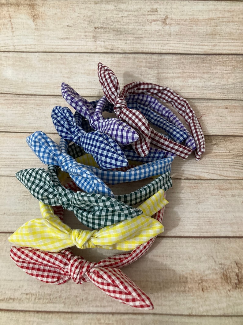 May include: A stack of six gingham fabric headbands in various colors, including red, yellow, blue, green, and purple. The headbands are tied in a bow at the top.