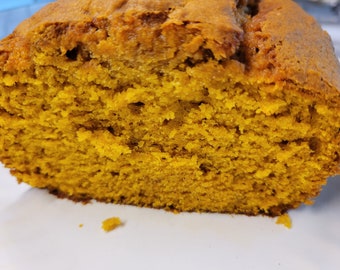 May include: A close-up of a slice of pumpkin bread. The bread has a golden-orange color and a moist, textured crumb. The crust is a darker shade of brown. The bread is on a white surface.