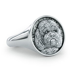 May include: A silver ring with a round, raised image of a dog's face. The dog has a fluffy, white coat and a serious expression.