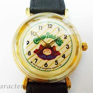 Puede incluir: Un reloj de pulsera dorado con una esfera blanca y una correa de cuero negra. La esfera presenta un logotipo de Cabbage Patch Kids y un fondo a rayas. El reloj tiene agujas negras y números dorados.