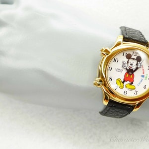 May include: Gold-tone Mickey Mouse watch with a black leather band. The watch face features a white background with Mickey Mouse in the center. The watch has a chime and alarm function.