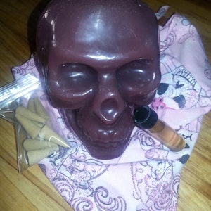 May include: A dark purple skull candle with a white wick, a small bag of incense cones, and a bottle of essential oil. The items are arranged on a pink and white patterned fabric.