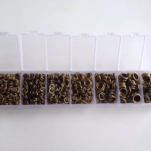 May include: A clear plastic storage box with eight compartments filled with gold jump rings. The jump rings are various sizes.