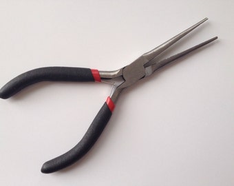 May include: Needle-nose pliers with black handles and red accents. The pliers are made of metal and have long, tapered jaws. The handles are textured for grip. The tool is designed for precision work.