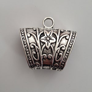 May include: Silver-toned metal pendant with an ornate, openwork design. The pendant features a central floral motif and scrolling patterns. It has a loop at the top for attaching to a chain or cord. The pendant has a slightly trapezoidal shape.