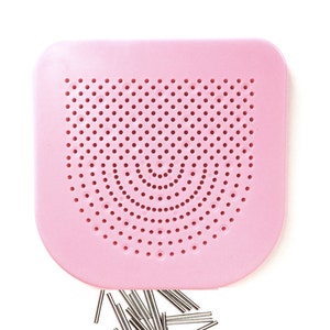 May include: A pink plastic pegboard with a circular pattern of holes. A pile of silver metal pegs are scattered below the board.