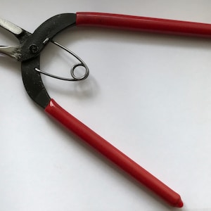 May include: Red handled pliers with black metal jaws. The pliers are open and a small metal ring is attached to the jaws.