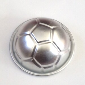 May include: A silver metal soccer ball shaped cake pan. The pan has a raised design of a soccer ball.