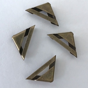 May include: Four antique brass-colored metal corner protectors. Each triangular piece has a diagonal black stripe and a cut-out section. These decorative accents are designed to protect the corners of books, boxes, or other items.