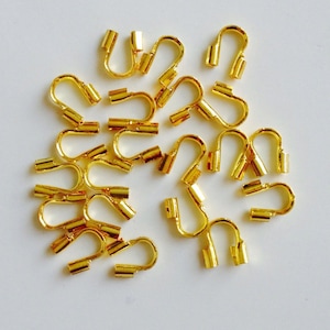 May include: A collection of small, gold-colored crimp end findings, also known as cord ends, for jewelry making. These findings have a U-shaped design with a flat, rectangular base and are scattered on a white surface.