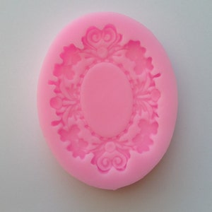 May include: Pink silicone mold for baking or crafting. The mold features an oval frame with a decorative floral design.