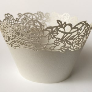 May include: A white paper cupcake liner with a decorative floral lace design around the top edge. The liner is cone-shaped and appears to be made of a sturdy paper material. The intricate cut-out pattern adds an elegant touch.