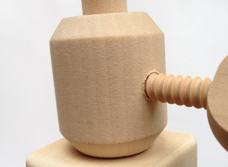 May include: Close-up of a wooden clamp with a screw mechanism. The clamp is made of light-colored wood and features a cylindrical body with a threaded screw extending from the side. The design is simple and functional, with a focus on the wood grain.