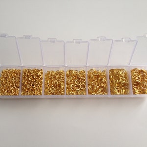 May include: A clear plastic storage box with eight compartments filled with gold jump rings. The jump rings are various sizes.