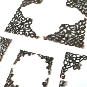 May include: A collection of bronze-coloured metal corner brackets with intricate filigree designs. The decorative elements feature swirling and floral patterns, perfect for adding a vintage aesthetic to craft projects. Measurements are in centimetres.