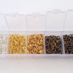May include: A clear plastic storage box with six compartments filled with silver, gold, and bronze jump rings. The box is open and the jump rings are visible.